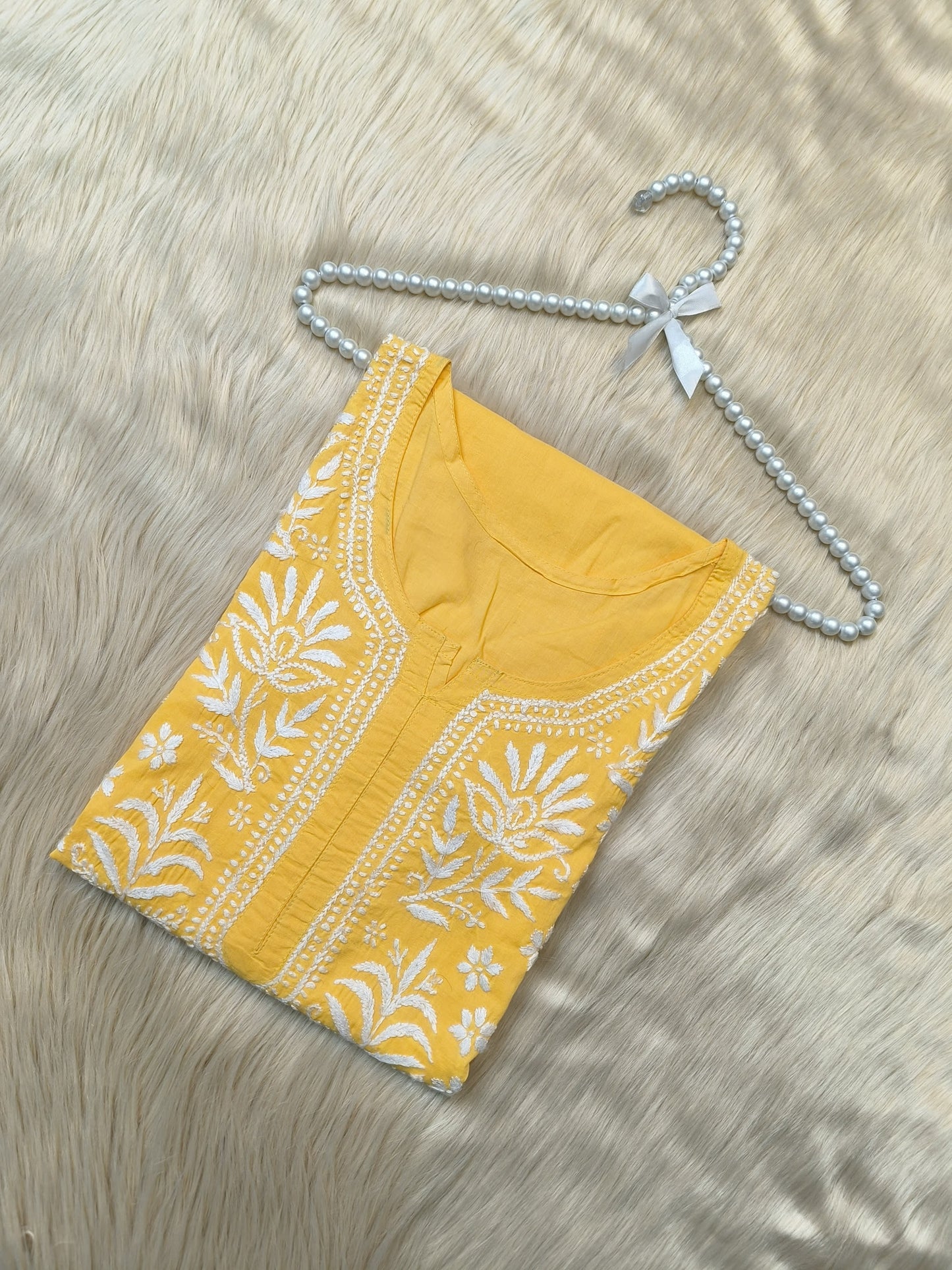 Yellow cotton kurta with white Lucknowi Chikankari hand embroidery displayed flat on fur background with pearl hanger